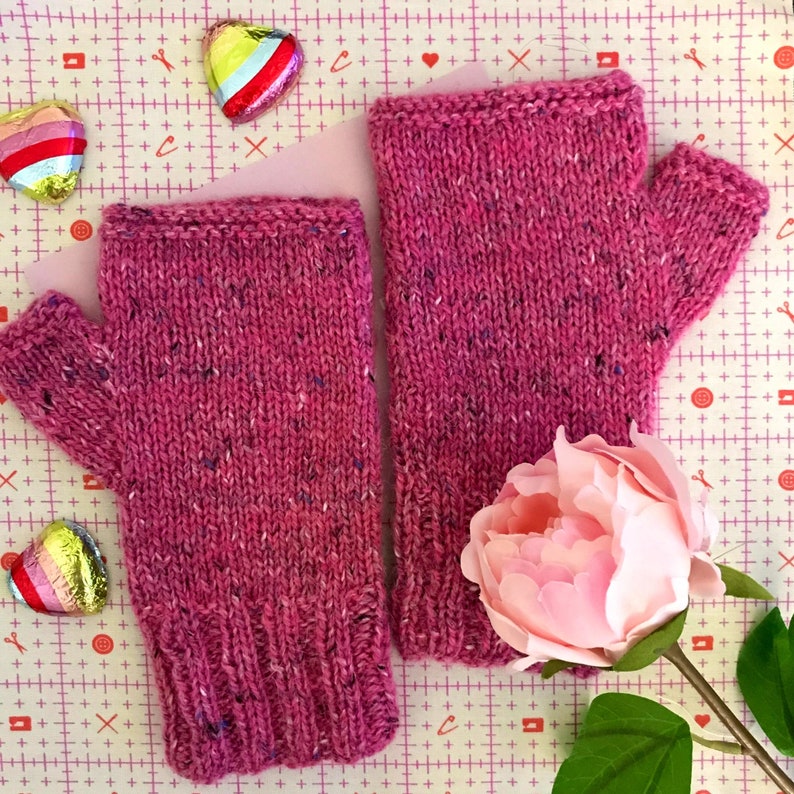May include: A pair of pink knitted fingerless gloves with a speckled pattern. The gloves are laying on a pink and white patterned surface with a pink flower.