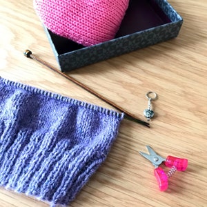 May include: Purple knitted fabric on knitting needles with a pink knitted hat in a box, a stitch marker, and pink scissors.
