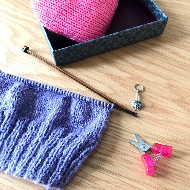 May include: Purple knitted fabric on knitting needles with a pink knitted hat in a box, a stitch marker, and pink scissors.