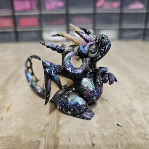 May include: A small dragon figurine featuring a dark navy and black body with a galaxy-inspired pattern of white stars, purple, and teal nebula clouds. The creature has curved horns, textured wings, and a long, curled tail with visible circular markings.