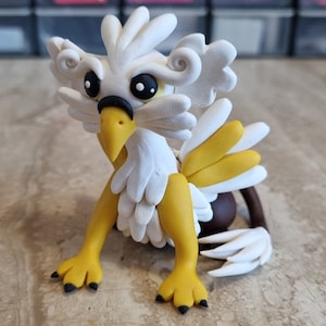 May include: A white and yellow clay sculpture of a griffin with black eyes and claws. The griffin is sitting on a light brown surface.
