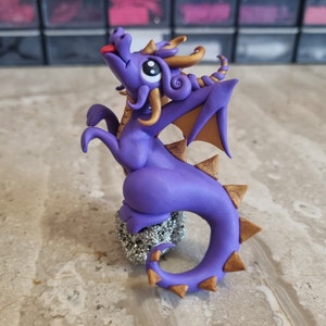 May include: Purple and gold dragon figurine with a spiky tail and wings. The dragon is sitting on a rock and has its tongue sticking out.