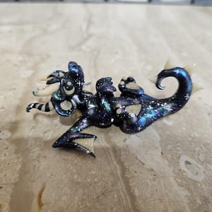 May include: A small, sculpted dragon figurine featuring a dark blue and black galaxy-inspired finish with white star-like speckles. The creature has cream-colored horns, wing tips, and tail spikes, with a curled tail and visible paw pads.