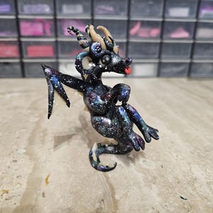 May include: A small, sculpted dragon figurine featuring a dark, galaxy-inspired paint finish with blue, purple, and white star-like speckles. The creature has curved tan horns, large black eyes, a small red tongue, and translucent wings with glitter.