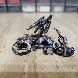 May include: A small dragon figurine featuring a black body with a galaxy-inspired paint pattern of white stars, purple, and blue nebula clouds. The creature has cream-colored spikes along its back, curved horns, and large eyes.