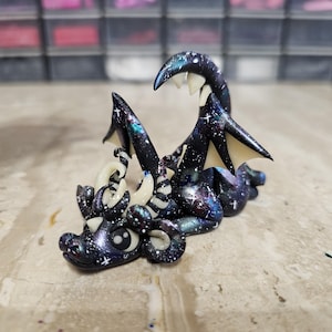 May include: A small dragon figurine featuring a dark galaxy-themed body with purple, blue, and white star-like speckles. The creature has cream-colored horns and wing membranes, large black eyes, and a curled tail, all crafted with a smooth finish.