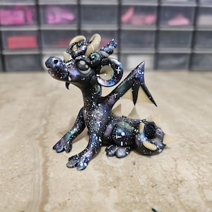 May include: A small dragon figurine featuring a dark galaxy-themed paint job with purple, blue, and white speckles resembling stars. The creature has cream-colored horns, wing accents, and tail spikes, with large eyes and a seated posture.