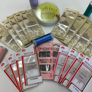 May include: Assortment of sewing needles in various sizes and types, including gold-eye crewel embroidery needles, knitter's needles, and household needles. The needles are packaged in clear plastic and labeled with their type and size. There are also spools of pink, blue, and green thread.