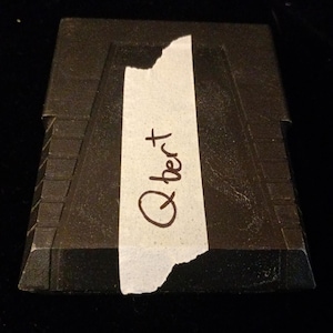 May include: A black, rectangular, pyramid-shaped object with a stepped, tiered design on the sides. A strip of white masking tape is applied vertically across the centre, featuring the handwritten text Q*bert in black ink.