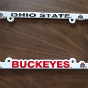 May include: A white rectangular license plate frame featuring the text OHIO STATE in black lettering at the top and BUCKEYES in red lettering at the bottom. The frame includes four small Ohio State University logos at each corner.