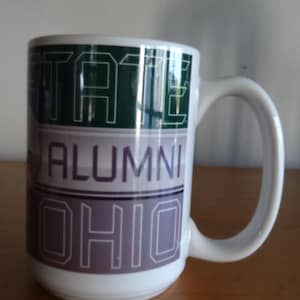 May include: White ceramic mug with the words "STATE ALUMNI OHIO" in green, purple, and white. The mug has a large, curved handle and is set against a neutral background.