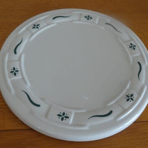 May include: A white ceramic plate with a decorative border featuring small green floral designs and curved lines. The plate is round and sits on a wooden surface. This plate is suitable for serving food.