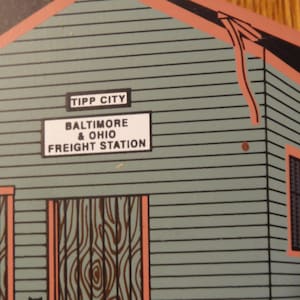 May include: Illustration of a vintage freight station with the text "TIPP CITY" and "BALTIMORE & OHIO FREIGHT STATION". The building is painted in shades of gray and has orange trim around the windows and doors. The wooden doors have a wood grain design.