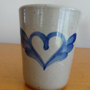 May include: A cylindrical ceramic tumbler with a speckled light gray finish. The exterior features a hand-painted blue heart design with stylized wing-like accents extending from the sides. The vessel has a smooth, glossy glaze and a simple, straight-sided profile.