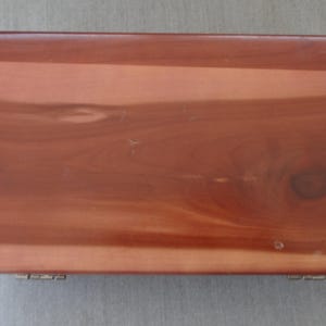 May include: A rectangular wooden box with a reddish-brown finish. The wood grain is visible, and the box has a glossy surface. The box is closed and has two small hinges on the front edge. The box appears to be made of cedar.