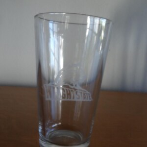 May include: A clear glass pint glass with the words "WRIGHT STATE" in white lettering. The glass has a slightly tapered shape and is empty. The glass is sitting on a wooden surface.