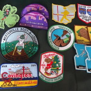 May include: A collection of twelve embroidered fabric patches featuring various shapes and designs, including a green clover, a riverboat, a bear decorating a tree, and text such as Westerville Boot-N-Leggers, Ohio Trekkers, and Sister Cities.