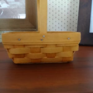 May include: A small, rectangular woven basket with a tall, arched handle. The basket is a light yellow color and has a woven design. The handle is attached to the basket with small metal rivets. The basket is sitting on a dark brown surface.