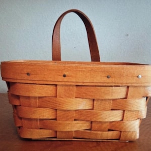 May include: A rectangular, woven wooden basket with a brown leather handle. The basket is made of light brown wood strips, with a darker brown handle. The basket is sitting on a brown surface.