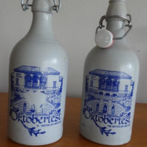 May include: Two light gray stoneware bottles with swing-top closures. Each bottle features a blue graphic of a building and the text "Oktoberfest" along with "The Dayton Art Institute" printed on the front surface of the ceramic body.