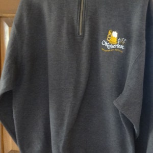 May include: A dark gray sweatshirt with a zipper up the front. The sweatshirt has an embroidered logo that says "Oktoberfest" with a beer stein graphic.