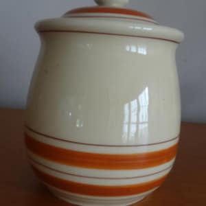 May include: A cream-colored ceramic canister with a matching lid, featuring horizontal orange stripes around the base and a single thin brown line near the top rim. The surface has a visible crackle glaze finish and a rounded, bulbous shape.
