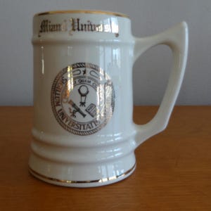 May include: Cream-colored ceramic mug with a gold rim and handle. The mug features the words "Miami University" in gold lettering and a gold seal with text and symbols. The mug is on a wooden surface.