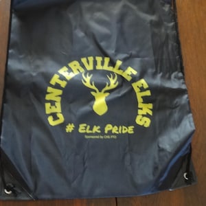 May include: A navy blue drawstring bag with the words "Centerville Elks" in a yellow arched font, with a yellow elk head logo. Below the logo is the text "# Elk Pride". The bag is on a wooden surface.