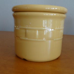 May include: A cylindrical yellow ceramic canister with a matching lid. The exterior features a raised, embossed basket-weave pattern around the upper section. The finish is smooth and glossy, reflecting light across the curved surface of the container.