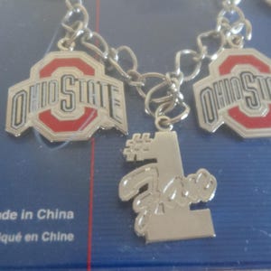May include: A silver charm bracelet with three charms. Two charms feature the Ohio State logo in red and black. The third charm is a silver number one with the word "Buckeye" written below it. The bracelet is made in China.