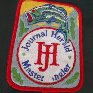 May include: A rectangular embroidered patch with a red border featuring a green fish above a white circular emblem. The emblem contains the red letters "HJ" and the blue text "Journal Herald" and "Master Angler" on a light blue background.
