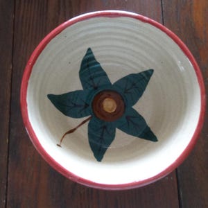 May include: A ceramic bowl with a cream-colored interior and a red rim. The bowl features a central design of a stylized, dark green and brown flower. The bowl is set on a dark wooden surface.