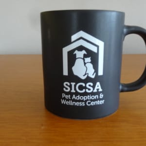 May include: Black ceramic coffee mug with a white logo that says "SICSA Pet Adoption & Wellness Center". The logo features a white silhouette of a house with a dog and cat inside.