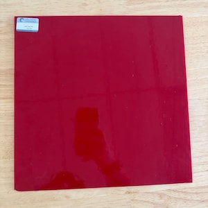May include: A square sheet of glossy red glass with a smooth, reflective surface. In the top left corner, there is a small rectangular white sticker with blue text that reads Bullseye Compatible and 250-125-FR Red Thin.