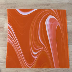 May include: A square sheet of acrylic material featuring a vibrant orange and white marbled pattern. The design consists of fluid, swirling lines of opaque white blended into a solid orange background, creating a smooth, glossy surface texture.