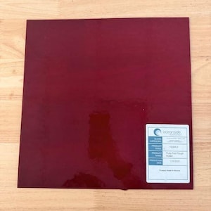May include: A square sheet of deep red glass with a smooth, reflective surface. A white rectangular label is affixed to the bottom right corner, featuring the Oceanside Glass & Tile logo, product code 110RR-F, and the name Ruddy Red Rough Rolled.