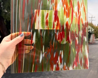 Puede incluir: Una lámina cuadrada de vidrio translúcido con un patrón decorativo de rayas verticales y formas orgánicas en tonos verde oliva, rojo brillante y blanco. El vidrio tiene un acabado liso y brillante con una etiqueta rectangular en el centro.