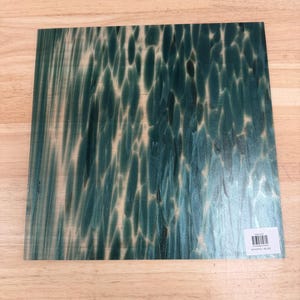 May include: A square sheet of glass featuring a dark teal and forest green pattern with elongated, vertical, organic shapes. The surface has a smooth, reflective finish and includes a small white rectangular sticker with a barcode in the corner.