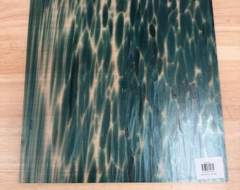 Puede incluir: Una lámina de vidrio cuadrada con un patrón de color verde bosque y verde azulado oscuro con formas orgánicas verticales alargadas. La superficie tiene un acabado liso y reflectante e incluye una pequeña etiqueta rectangular blanca con un código de barras.