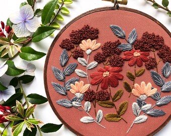 May include: A circular wooden embroidery hoop containing a dusty rose fabric background with hand-stitched floral designs. The embroidery features textured rust-colored clusters, deep red flowers, pale peach blossoms, and grey-blue serrated leaves.