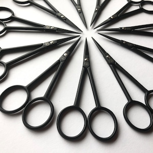 Può includere: Una collezione di forbici in metallo nero opaco disposte in un motivo circolare. Ogni paio di forbici presenta lame sottili e appuntite con anelli circolari, e una piccola vite color argento che fissa le due lame nel punto di articolazione.