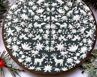 Puede incluir: Un bastidor de bordado circular con hilo blanco cosido sobre tela verde oscuro. El diseño muestra un patrón simétrico de flores blancas, hojas, zorros, ciervos y pájaros dispuestos en un motivo botánico intrincado y repetitivo.