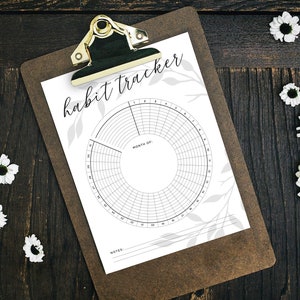 May include: A black and white habit tracker on a clipboard with a gold clip. The tracker has a circle divided into 31 sections with a smaller circle in the center. The text "habit tracker" is written at the top of the page.