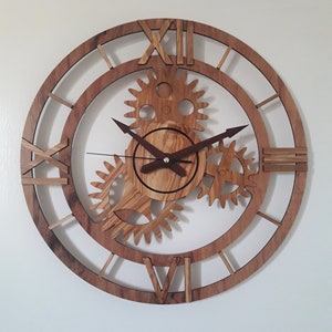 May include: A wooden wall clock with a gear design. The clock features Roman numerals and a circular frame. The gears and hands are a darker wood tone, contrasting with the lighter wood of the frame. The clock is mounted on a white wall.