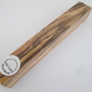 May include: A rectangular piece of olive wood with a natural wood grain pattern. The wood is light brown with dark brown streaks. The wood is labeled "Holly Land Olive Wood" and "Made in Bethlehem".