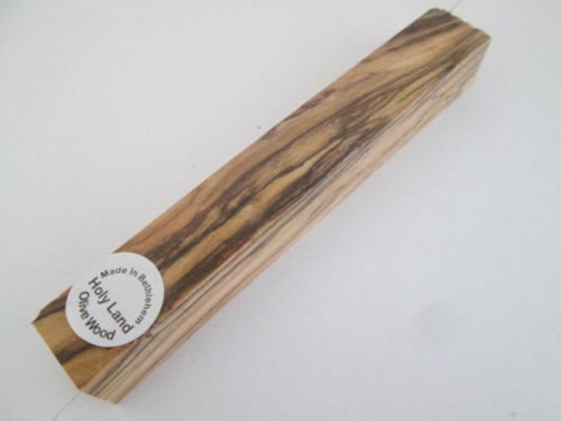 May include: A rectangular piece of olive wood with a natural wood grain pattern. The wood is light brown with dark brown streaks. The wood is labeled "Holly Land Olive Wood" and "Made in Bethlehem".