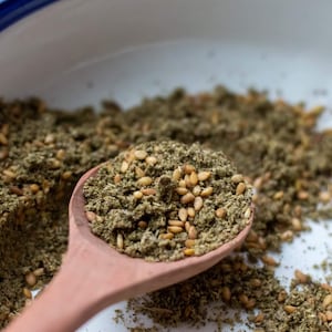 May include: A wooden spoon filled with a blend of spices and sesame seeds, set against a backdrop of the same mixture in a white bowl with a blue rim. The spices appear to be a mix of green and brown.