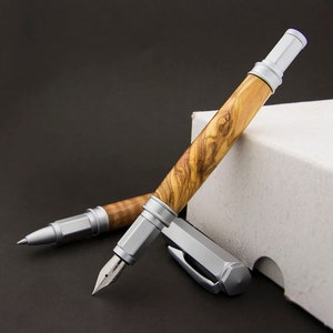 May include: Two pens with silver accents and wooden barrels. The pens are made of olive wood with a natural grain pattern. The pens are lying on a white box.