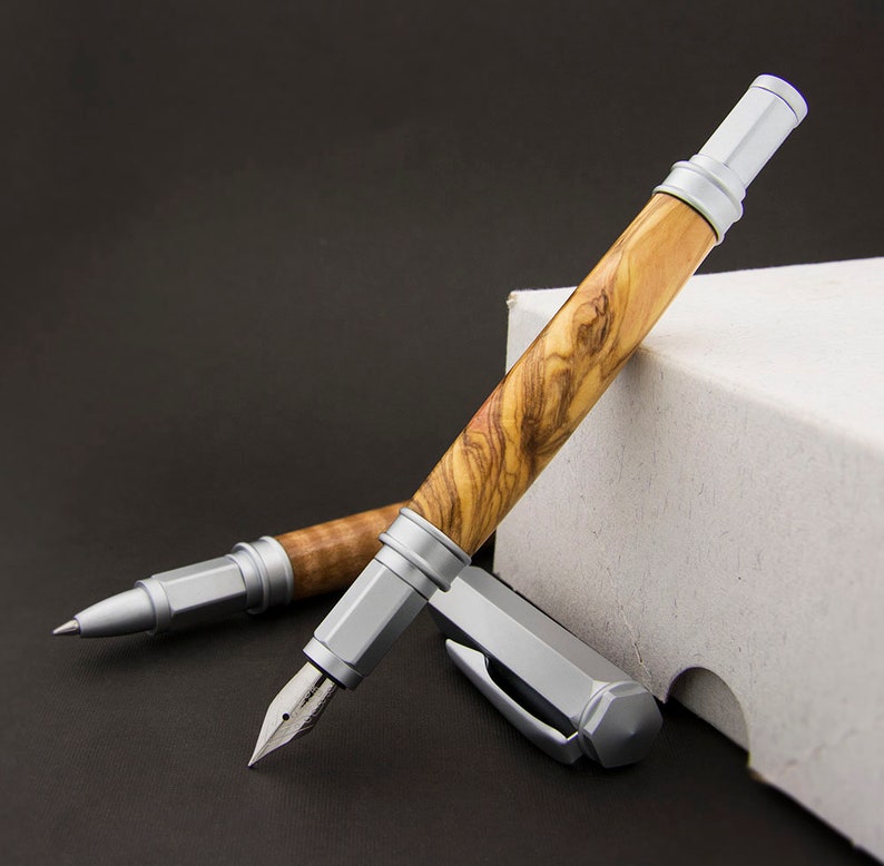 May include: Two pens with silver accents and wooden barrels. The pens are made of olive wood with a natural grain pattern. The pens are lying on a white box.