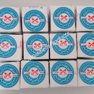 Puede incluir: Doce cajas blancas con etiquetas circulares azules y rojas. Cada etiqueta tiene texto árabe y una imagen roja de dos llaves cruzadas. El texto en la etiqueta dice "صابون النابلسي الممتاز" que se traduce como "Jabón Superior de Nablus".